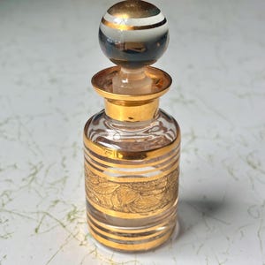 May include: A clear glass perfume bottle with gold accents and a decorative gold leaf pattern. The bottle has a gold-colored stopper with a striped, spherical top. The bottle is approximately 4 inches tall.