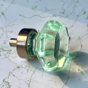 May include: A light green, faceted glass cabinet knob with a brass base and stem. The knob has a geometric shape and is designed for furniture. The brass hardware adds a touch of vintage charm.