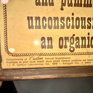 May include: Vintage framed advertisement with text reading "unconsciousness" and "an organic ca." The frame is brown, and the background is a light tan color. The text "Compliments of Carlson Natural Supplements" is also visible.