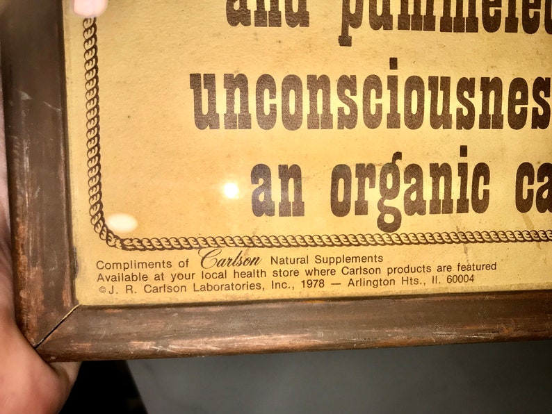 May include: Vintage framed advertisement with text reading "unconsciousness" and "an organic ca." The frame is brown, and the background is a light tan color. The text "Compliments of Carlson Natural Supplements" is also visible.