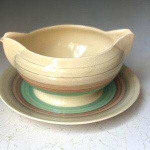Puede incluir: Una salsera vintage de color crema con un platillo a juego. El recipiente tiene una forma única con un pico y asas. El platillo y el recipiente están decorados con rayas horizontales en tonos marrón, verde y crema.