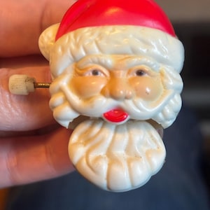 May include: A wind-up Santa Claus toy featuring a bright red hat, a white moulded beard and moustache, and a small white winding key on the side. The face has painted brown eyes, rosy cheeks, and red lips, with a separate piece for the lower beard.