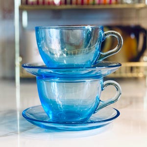 Può includere: Due tazze da tè impilate in vetro blu trasparente con piattini abbinati. Le tazze hanno manici curvi e poggiano su una superficie bianca. Lo sfondo include una mensola color oro e altri oggetti da cucina.
