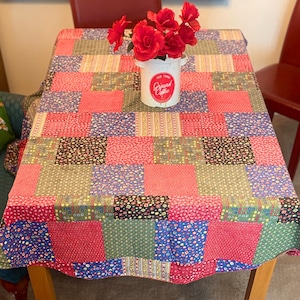 May include: A rectangular patchwork tablecloth featuring a variety of floral and geometric patterns in shades of red, blue, green, and yellow. A white ceramic canister with the red text "Fair Trade Ground Coffee" holds a bouquet of red flowers.