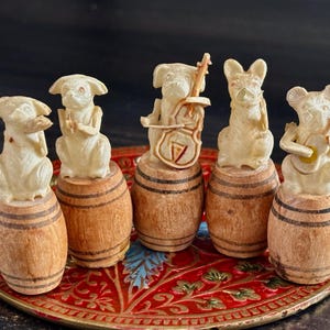 May include: Five off-white carved animal figurines, each perched atop a small wooden barrel with dark horizontal bands. The figures include animals playing musical instruments like a cello and guitar, while others appear to be singing or clapping.
