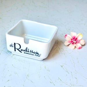 May include: A square white ceramic ashtray with a notched rim. The front features black printed text that reads the Radisson MINNEAPOLIS hotel. The ashtray has a smooth, glossy finish and a clean, simple design suitable for a tabletop display.
