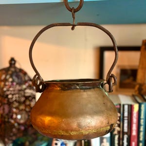 May include: A vintage copper pot with a hanging loop and a rustic iron handle. The pot is hanging from a shelf and has a worn, aged appearance.
