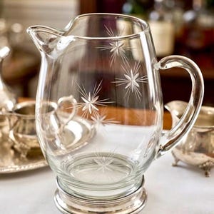 Puede incluir: Una jarra de cristal transparente con un patrón de estrellas grabado en el cuerpo. La jarra tiene un asa curva, un pico vertedor y una base de metal pulido en tono plateado. El diseño incluye varios grabados en forma de estrella en el cristal.