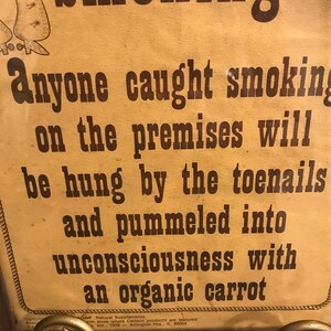 May include: A vintage sign with the word "Smoking" at the top. The sign warns that anyone caught smoking on the premises will be punished. The text is in brown ink on a tan background. The sign is held by a decorative metal stand.
