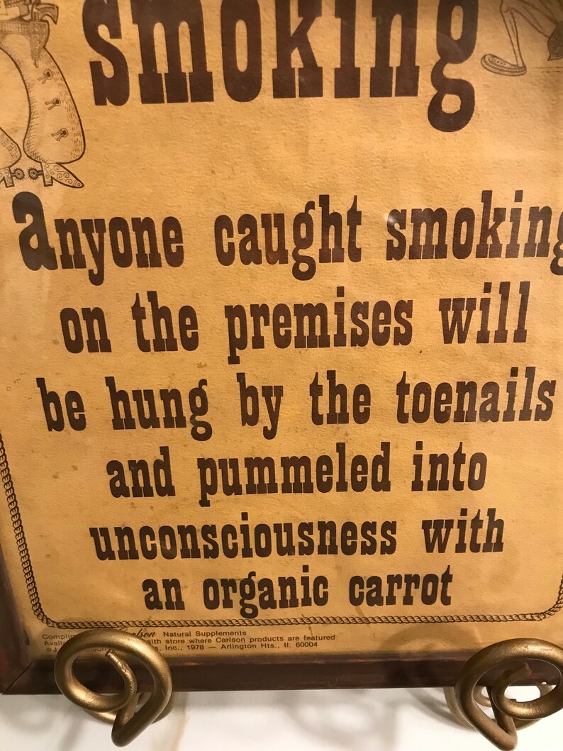 May include: A vintage sign with the word "Smoking" at the top. The sign warns that anyone caught smoking on the premises will be punished. The text is in brown ink on a tan background. The sign is held by a decorative metal stand.