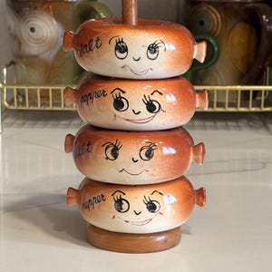 Pode incluir: Uma pilha de quatro saleiros e pimenteiros de cerâmica com um design de rosto sorridente. Os saleiros e pimenteiros são laranja e castanhos com as palavras "sal" e "pimenta" escritas neles. Os saleiros e pimenteiros são empilhados numa base de madeira.