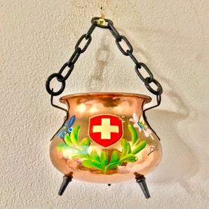 Könnte beinhalten: Ein hängender Kupfertopf mit Schweizer Flagge und Blumenmuster. Der Topf hat drei Beine und hängt an einer schwarzen Kette.