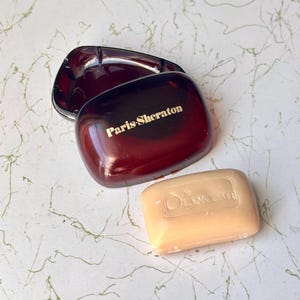 Puede incluir: Un jabonera marrón con el texto "Paris-Sheraton" en letras doradas, junto a una pastilla de jabón beige claro. La jabonera está abierta, revelando un interior brillante. El jabón tiene el texto "de Lancome" en relieve.