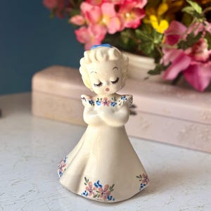 Peut inclure: Une figurine d'ange en porcelaine blanche aux yeux fermés, portant un nœud bleu et une robe à fleurs roses et bleues.