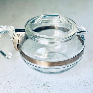 Puede incluir: Una tetera de vidrio transparente con una banda plateada en el medio y un asa transparente. La tapa también es de vidrio transparente. La tetera tiene un pico para verter. La tetera está sobre una superficie blanca.