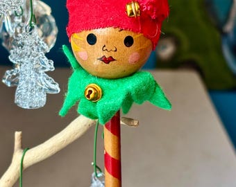 May include: A whimsical Christmas ornament shaped like an elf. The elf has a wooden head with painted features, a red felt hat, and a green felt collar. The body is a red and gold striped stick with a green ball at the bottom.