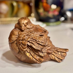 May include: A bronze-colored bird figurine with detailed wing and feather textures. The sculpture is in a resting pose, with its head tucked down. The bird is made of a solid material and has a metallic finish, suggesting it is a decorative item.