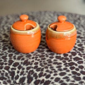 May include: Two small, orange ceramic jars with matching lids. Each jar has a small handle on the lid and a light brown band around the top. The jars are sitting on a leopard print surface.