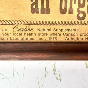 May include: A framed advertisement with the text "an organic" in large brown letters. The bottom of the frame includes text that reads "Compliments of Carlson Natural Supplements". The frame is dark brown.