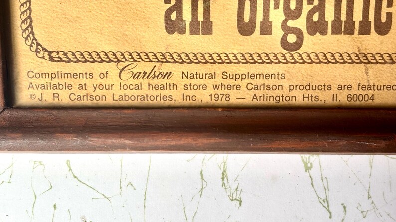 May include: A framed advertisement with the text "an organic" in large brown letters. The bottom of the frame includes text that reads "Compliments of Carlson Natural Supplements". The frame is dark brown.