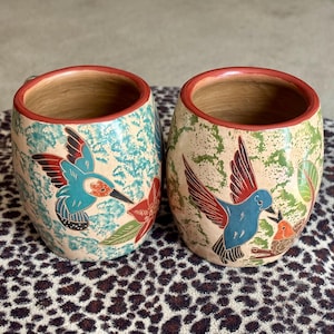 May include: Two ceramic mugs with a rounded shape and a red rim. The left mug features a turquoise sponge-painted background with a hummingbird and red flower. The right mug has a green sponge-painted background with a blue hummingbird.
