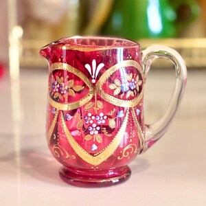 Könnte beinhalten: Ein kranberryroter Glaskrug mit verzierten goldfarbenen Bögen und floralen Motiven in Weiß und Blau. Das Gefäß verfügt über einen Henkel aus klarem Glas, einen kleinen Ausguss und einen runden Fuß mit detaillierten, handgemalten Verzierungen.