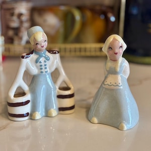 Könnte beinhalten: Zwei Keramikfiguren: eine Frau in blauer und weißer Uniform, die Eimer trägt, und eine Frau in blauem Kleid mit weißer Schürze. Beide haben blondes Haar und rosige Wangen. Die Figuren sind wahrscheinlich Vintage oder Sammlerstücke.