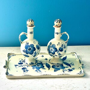 May include: Two ceramic cruets with spherical bodies, handles, and spherical stoppers, decorated with blue floral patterns on a cream background. One cruet is marked with the letter O and the other with A, both resting on a matching rectangular tray.