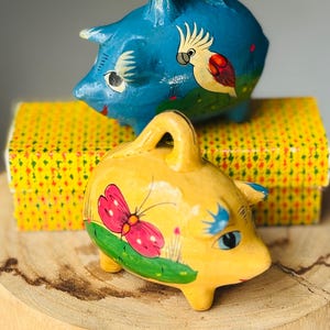 May include: Two small piggy banks, one blue and one yellow, each featuring a handle on top and hand-painted details. The blue bank displays a white cockatoo, while the yellow bank features a pink butterfly. Both have painted eyes and green grass.