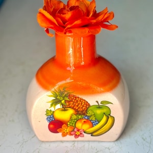 May include: An orange ceramic vase with a white base, featuring a colorful fruit design. The vase is topped with a vibrant orange flower. The fruit design includes a pineapple, apples, bananas, and grapes.