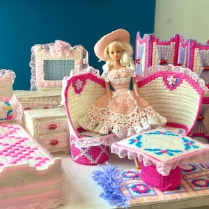 May include: A doll wearing a pink dress and hat sits in a miniature crocheted chair. The scene includes a bed, table, and other furniture, all crafted with pink, white, and blue yarn, creating a charming dollhouse setting.
