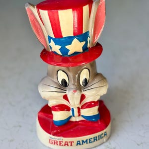Può includere: Statuetta in ceramica di Bugs Bunny con cappello e papillon a stelle e strisce. La base rossa riporta la scritta "Marriott's GREAT AMERICA". Il coniglio ha il pelo grigio e grandi occhi, un oggetto da collezione patriottico.