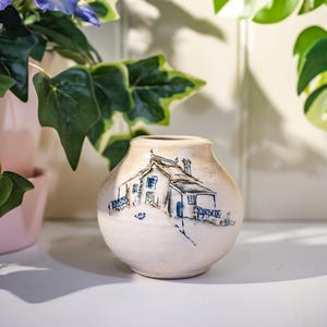 May include: A small, cream-coloured ceramic vase featuring a hand-painted house illustration. The house has a brown roof, blue shutters, and a blue door. The vase is round with a narrow neck. Decorative vase.