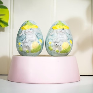 May include: Two ceramic Easter egg ornaments. Each egg is light blue with a white bunny wearing a yellow hat. The bunnies are surrounded by yellow flowers and green leaves. The eggs sit on a pink pedestal.