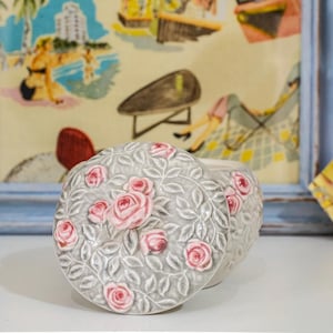 May include: A grey ceramic container with a lid, decorated with raised pink roses and leaves. The container sits on a white surface, with a framed picture in the background. The container is likely for storage or decoration.