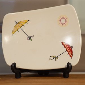 May include: A white ceramic rectangular serving dish with a yellow and red umbrella design. The dish has a sun design in the top right corner.
