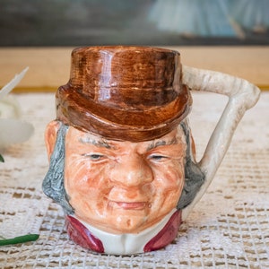 May include: A ceramic mug shaped like a man's head with a brown hat and a white handle. The mug has a detailed face with a moustache and beard.