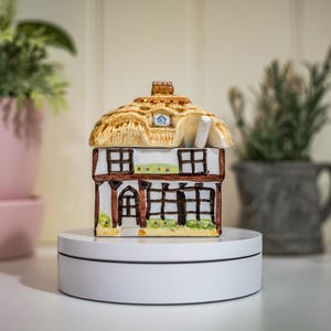 May include: A ceramic cottage-shaped container with a thatched roof, brown timber-framed walls, and small windows. The container is on a white rotating display stand, with potted plants in the background.