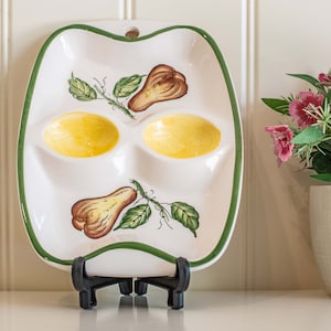Peut inclure: Plat de service en céramique blanche avec une bordure verte, comprenant deux puits jaunes en forme d'œuf et des illustrations de poires. Le plat est présenté sur un support noir, avec un petit trou en haut pour accrocher. Décoratif et fonctionnel.