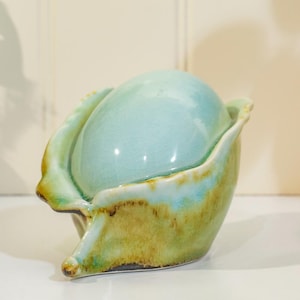 May include: A ceramic decorative bowl with a light blue, glossy finish. The bowl has a unique shape, with a rounded top and a flared, petal-like base. The base has a green and brown glaze, adding a touch of earthiness to the design.