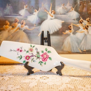 May include: White ceramic cake server with a floral design featuring pink and white flowers, green leaves, and a small bird. The server is on a black stand.