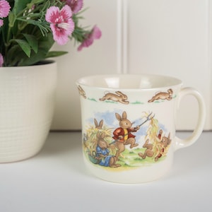 以下が含まれることがあります： ハンドルが付いた白いセラミック製のマグカップで、カラフルなウサギがフィールドにいるイラストが描かれています。ウサギは服を着て、道具を持っています。マグカップは白い背景に緑色の縁取りがされています。