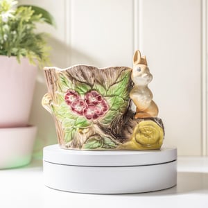 以下が含まれることがあります： 木株にウサギの置物がある陶器の植木鉢。植木鉢は茶色、緑、ブルゴーニュの花柄です。ウサギは薄茶色と白です。植木鉢は白い回転台にあります。