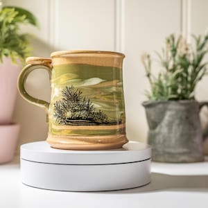 May include: A handmade ceramic mug with a landscape design. The mug features a green and tan colour scheme with a black tree illustration. The mug is approximately 10 cm tall and has a handle.