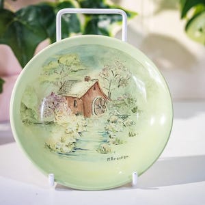 May include: A round, pale green decorative plate with a hand-painted landscape scene. The artwork shows a small brown house with a water wheel, surrounded by trees and a stream. The plate is held by a white metal stand.