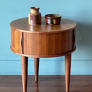 May include: A mid-century modern round side table made of wood, featuring a cylindrical body with a sliding door and three tapered legs. Two ceramic vessels with colorful vertical stripes sit on top. The table is approximately 24 inches in diameter.