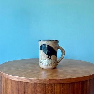 Puede incluir: Una taza de cerámica con un acabado moteado beige y un diseño de pájaro negro. La taza tiene un asa y un borde texturizado. El pájaro está posado sobre un diseño blanco texturizado. La taza está sobre una mesa de madera.
