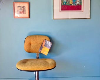 May include: A mustard yellow office chair with a rounded seat and back, set against a teal wall. The chair has a chrome base with wheels. Two framed artworks hang on the wall.