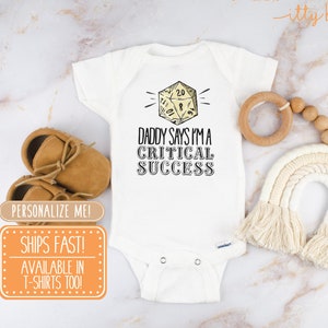 May include: White baby onesie with a yellow 20-sided die graphic and the text "Daddy says I'm a critical success".