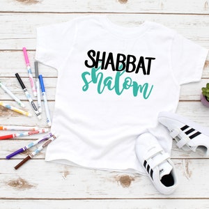 May include: White t-shirt with black and teal blue text that says "Shabbat Shalom".